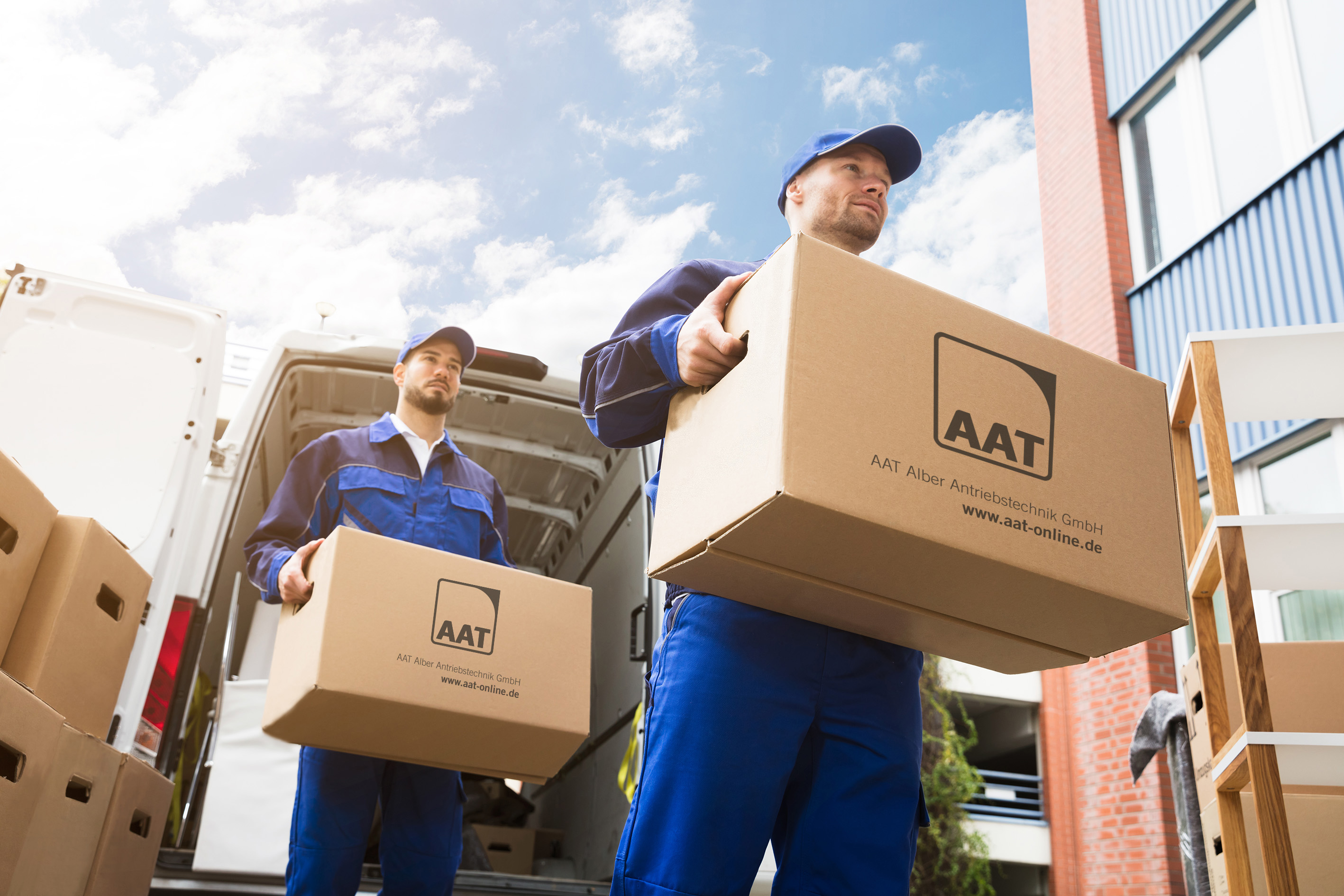 Das Bild zeigt zwei Männer eines Lieferdienstes, die zwei größere Pakete transportieren. Sie sehen dabei nicht gestresst aus. Das Bild steht symbolisch für reibungslöse Arbeitsabläufe ohne Verzögerungen in der Bearbeitung.