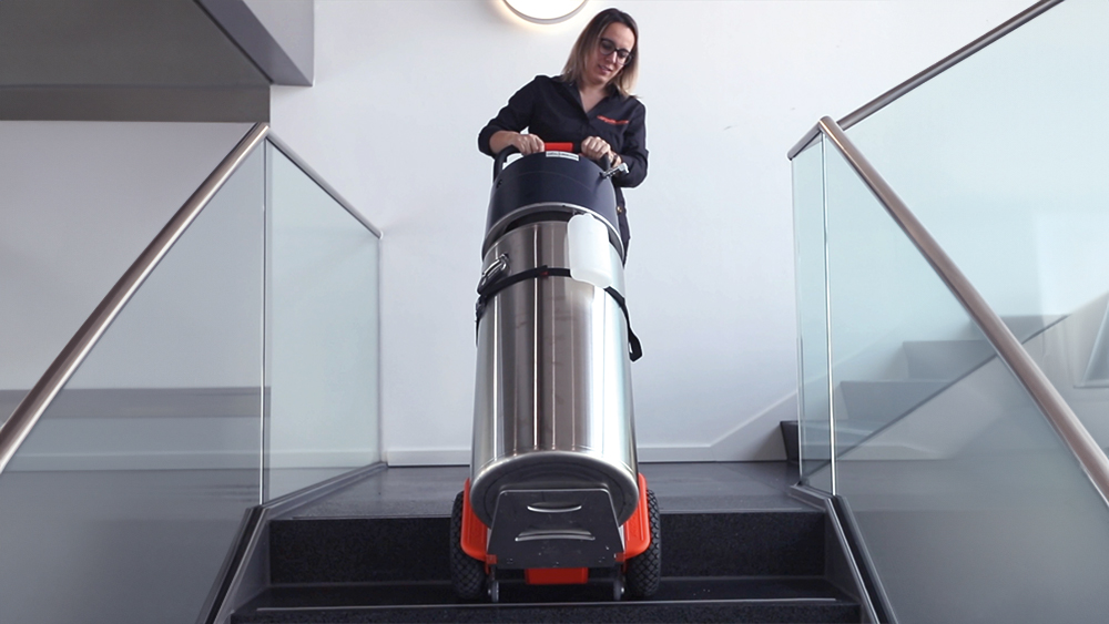 Das Bild zeigt eine Frau, die eine schwere Sauerstoffflasche mit Hilfe eines Lastentreppensteigers CargoMaster C120 von AAT über Treppen befördert.