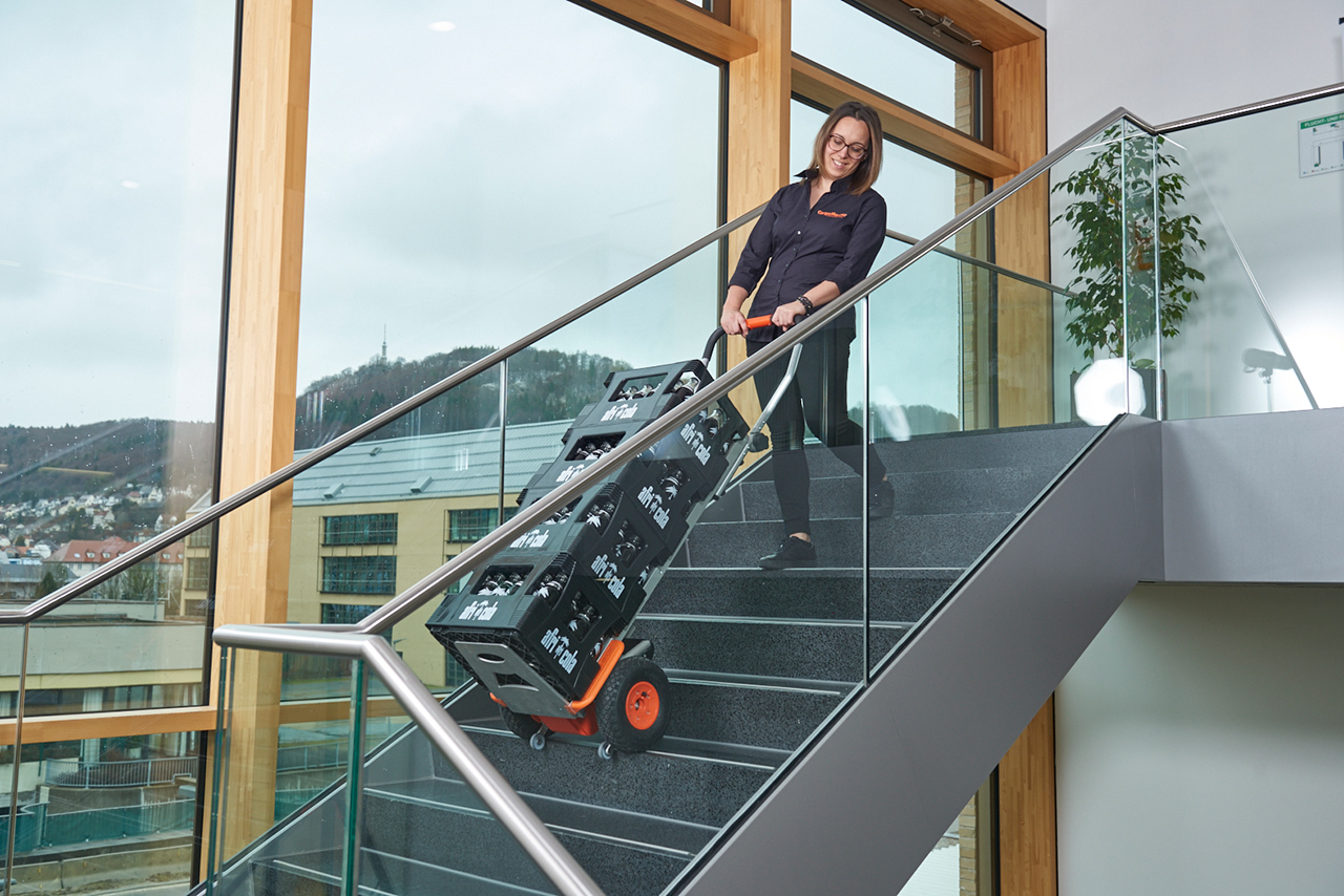 Das Bild zeigt eine Frau mittleren Alters, die mit Hilfe eines CargoMaster Treppensteiger für Lasten gleich fünf große, grüne Körbe mit Lebensmitteln auf einmal über Treppen befördert. Die Frau sieht dabei sehr entspannt aus und braucht kaum Kraft. 