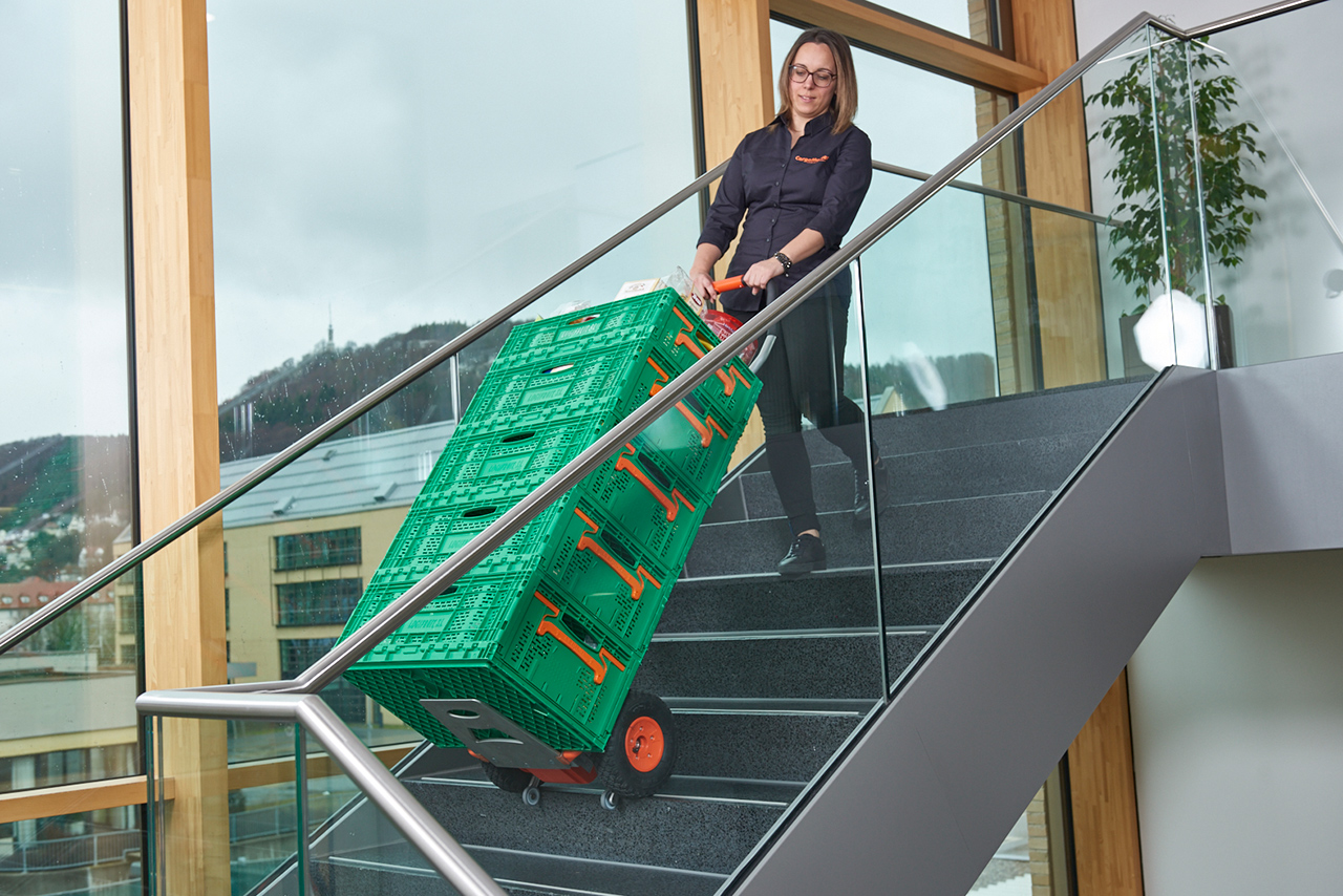 Das Bild zeigt eine Frau mittleren Alters, die mit Hilfe eines CargoMaster Treppensteiger für Lasten gleich fünf große, grüne Körbe mit Lebensmitteln auf einmal über Treppen befördert. Die Frau sieht dabei sehr entspannt aus und braucht kaum Kraft. 