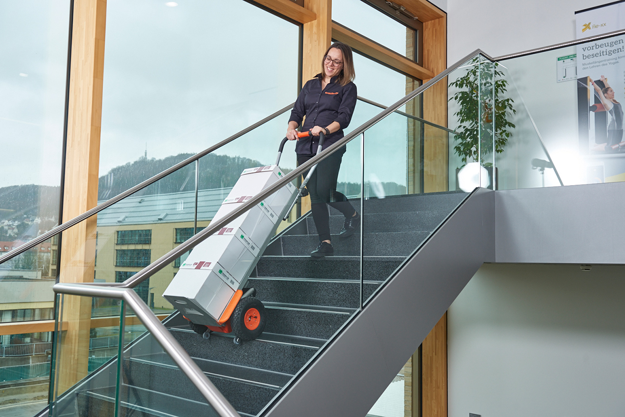 Das Bild zeigt eine Frau mittleren Alters, die mit Hilfe einer mobilen Treppensackkarre von AAT einen Stapel schwerer Papierkartons über Stufen befördert.