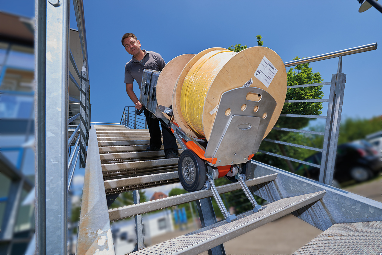 Auf dem Bild ist ein Mitarbeiter eines Handwerksunternehmens zu sehen, der mit einem Treppensteiger CargoMaster eine große, und schwere Rolle mit Material eine Außentreppe hinauf- oder hinab befördert. Er bedient den Treppensteiger alleine und braucht keine Unterstützung, um die schwere Last zu transportieren.