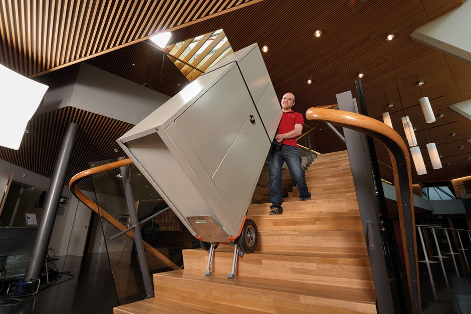 Das Bild zeigt einen Mann, der mit Hilfe eines CargoMaster Treppensteiger für Lasten, alleine, ohne Hilfe einen sehr großen Metallschrank über eine Treppe in den zweiten Stock transportiert.
