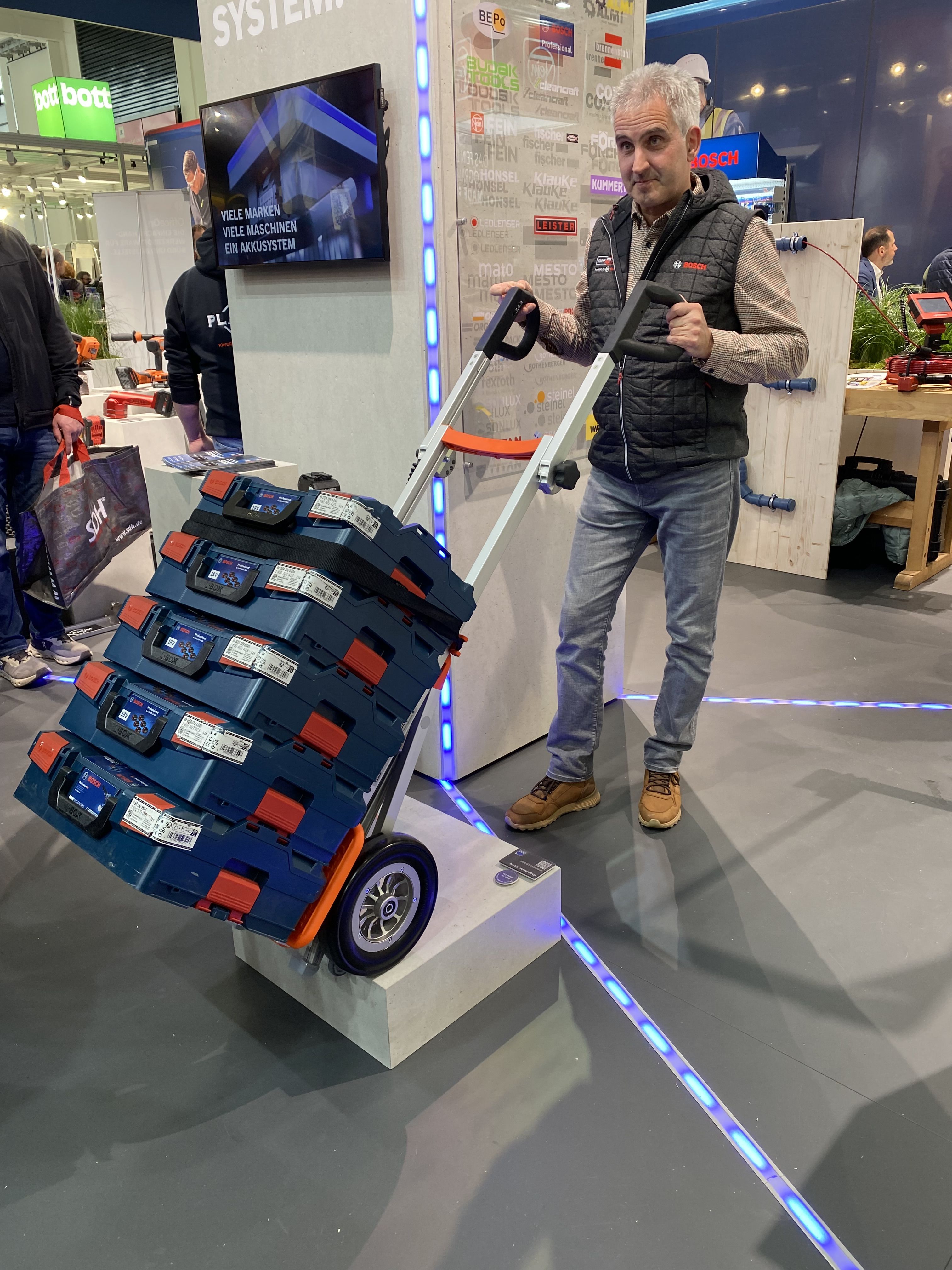 Das Bild zeigt einen Mann, der mit einem Treppensteiger eine große Maschine sicher eine Treppe hinauf transportiert, während im Hintergrund ein Gebäude mit Fenstern sichtbar ist. Rechts sticht ein orangefarbener Bereich mit einem Daumen-hoch-Symbol hervor, begleitet von der weißen Schrift: „BERATUNG & KOSTENLOSE VORFÜHRUNG“.