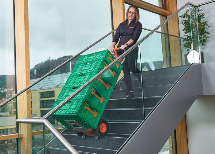 The picture shows a woman using a CargoMaster stair climber for loads to transport several boxes of groceries up a flight of stairs. You can also see the climbing legs of the stair climber and the safety brakes, which have stopped automatically at the edge of the stairs.