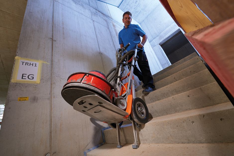 Das Bild zeigt einen Mann, der mit Hilfe eines CargoMaster Treppensteiger für Lasten, alleine, ohne Hilfe eine große Maschine über eine Treppe in den zweiten Stock transportiert.