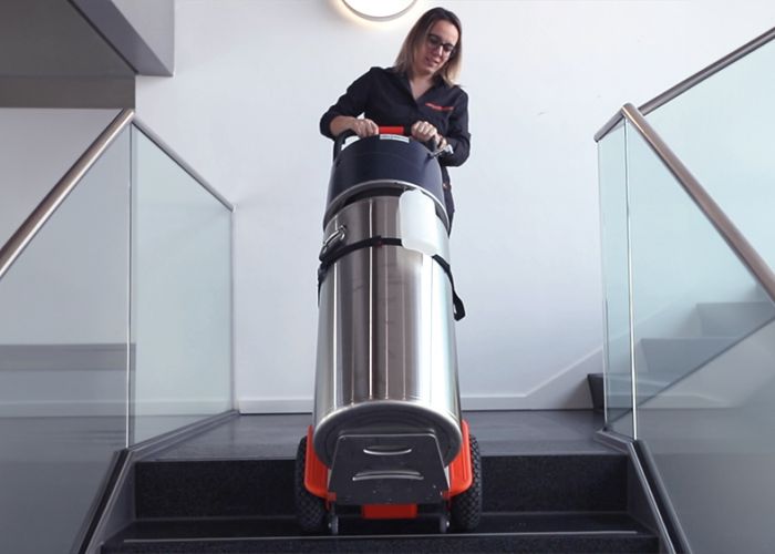 The picture shows a woman using a CargoMaster stair climber for loads to transport an oxygen cylinder up a flight of stairs. You can also see the climbing legs of the stair climber and the safety brakes, which have stopped automatically at the edge of the stairs.