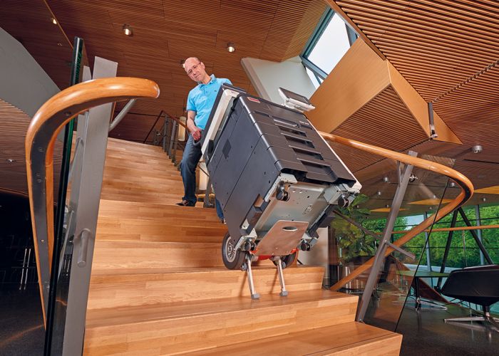 Op de foto wordt de CargoMaster trapklimmer gebruikt met een groot kopieerapparaat dat via een moderne houten trap in een gemeentehuis naar boven wordt getransporteerd.