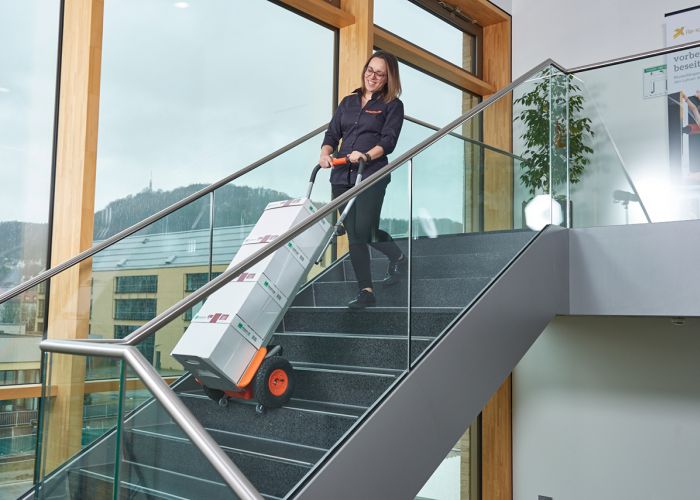 The picture shows a woman using a CargoMaster stair climber for loads to transport several boxes of copy paper up a flight of stairs. You can also see the climbing legs of the stair climber and the safety brakes, which have stopped automatically at the edge of the stairs.