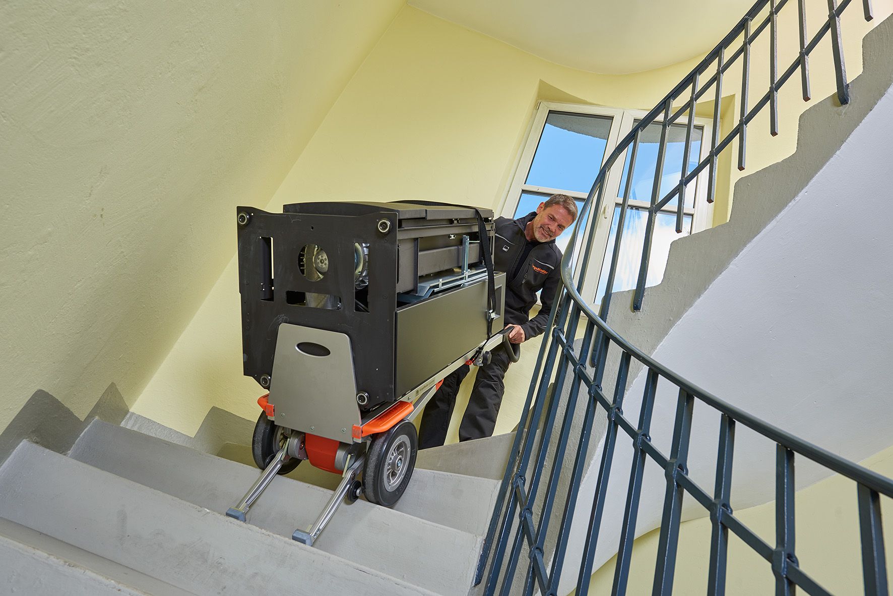 De foto toont een man van middelbare leeftijd die een zware automaat met behulp van een AAT CargoMaster C120 trappenklimmer een wenteltrap opdraagt.