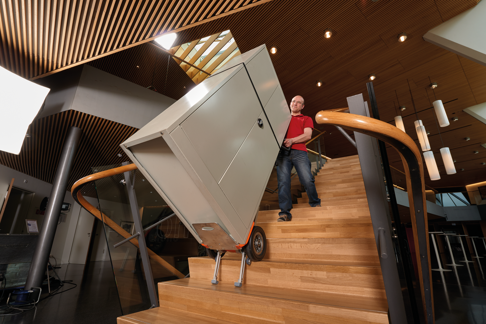 Een man van middelbare leeftijd vervoert alleen en zonder hulp een zware en volumineuze metalen kast een houten trap op met behulp van een CargoMaster trapklimmer van AAT, model CC160 Li-Ion. U kunt ook de verlengde klimpoten zien, die zachtjes op de trap neerkomen zonder deze te beschadigen.